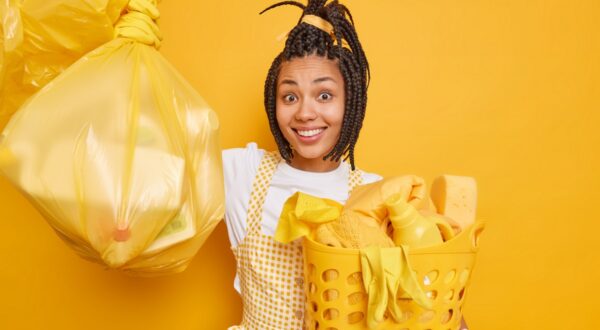 Smiling Afro American woman with dreadlocks enjoys housework holds polythene bag full of litter basket of laundry wears apron satisfied after productive cleaning poses against yellow studio wall